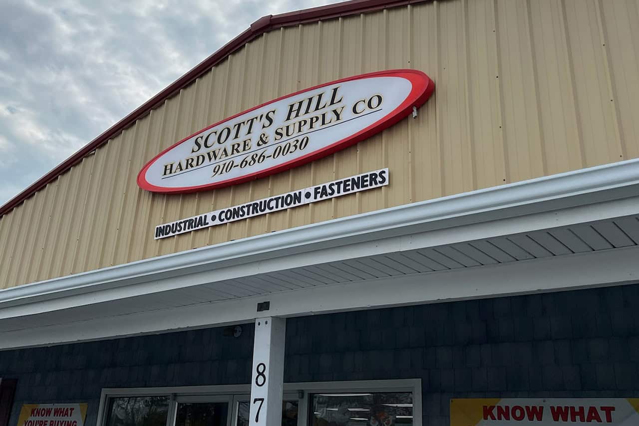 Exterior view of Scott's Hill Hardware & Supply Co., featuring their sign with contact number and services offered: industrial, construction, and fasteners. The building boasts a beige and blue facade, epitomizing the reliability and quality associated with Scott's Hill Hardware.
