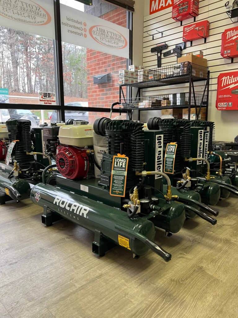 Several industrial air compressors on display in Scott's Hill Hardware. The green compressors are labeled "Rolair". The store also features various tools and equipment on shelves, with signage adorning the walls.