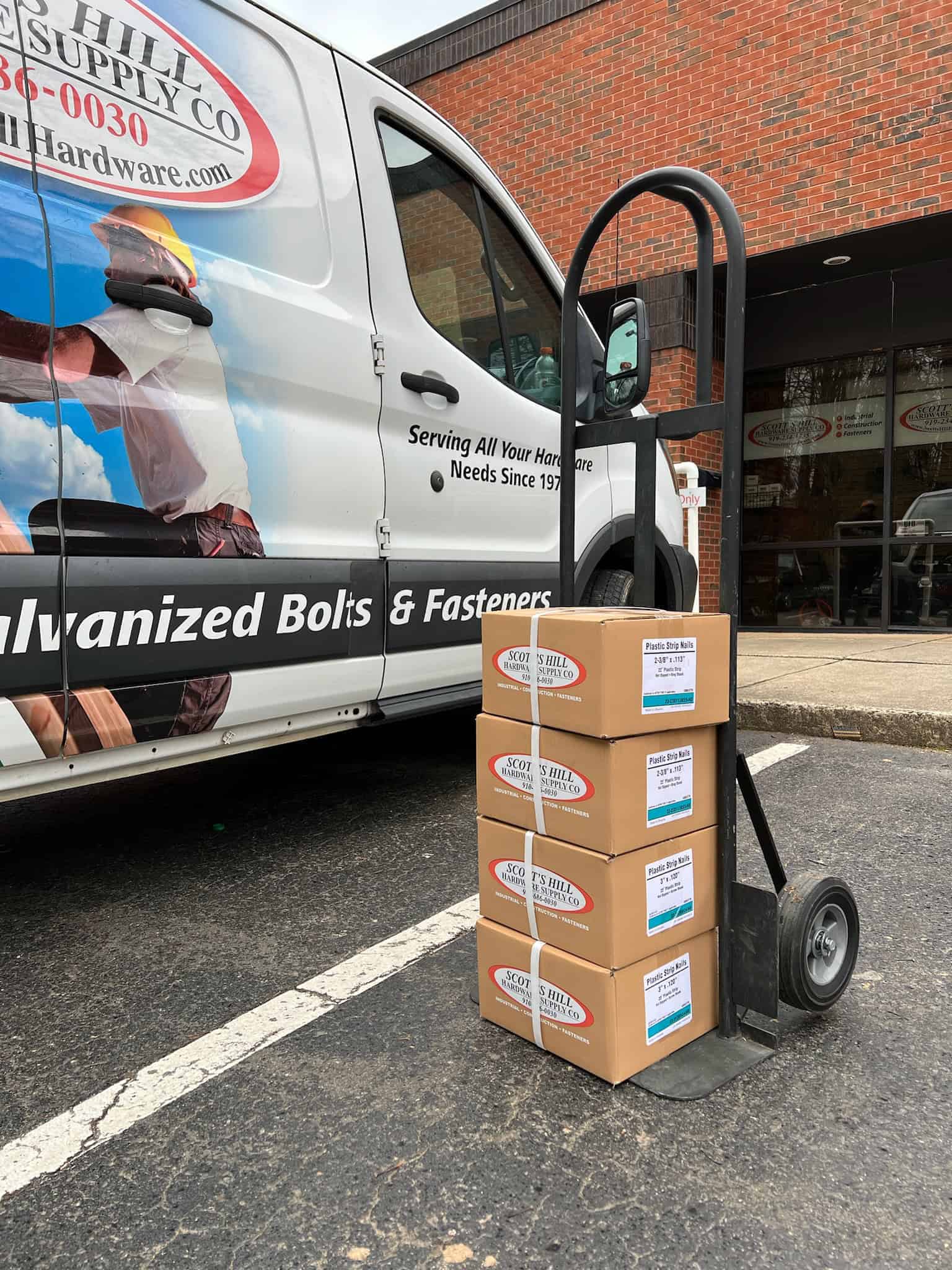 A dolly with four stacked boxes labeled "Hill Supply" sits in front of a Scott's Hill Hardware van parked beside a brick building.