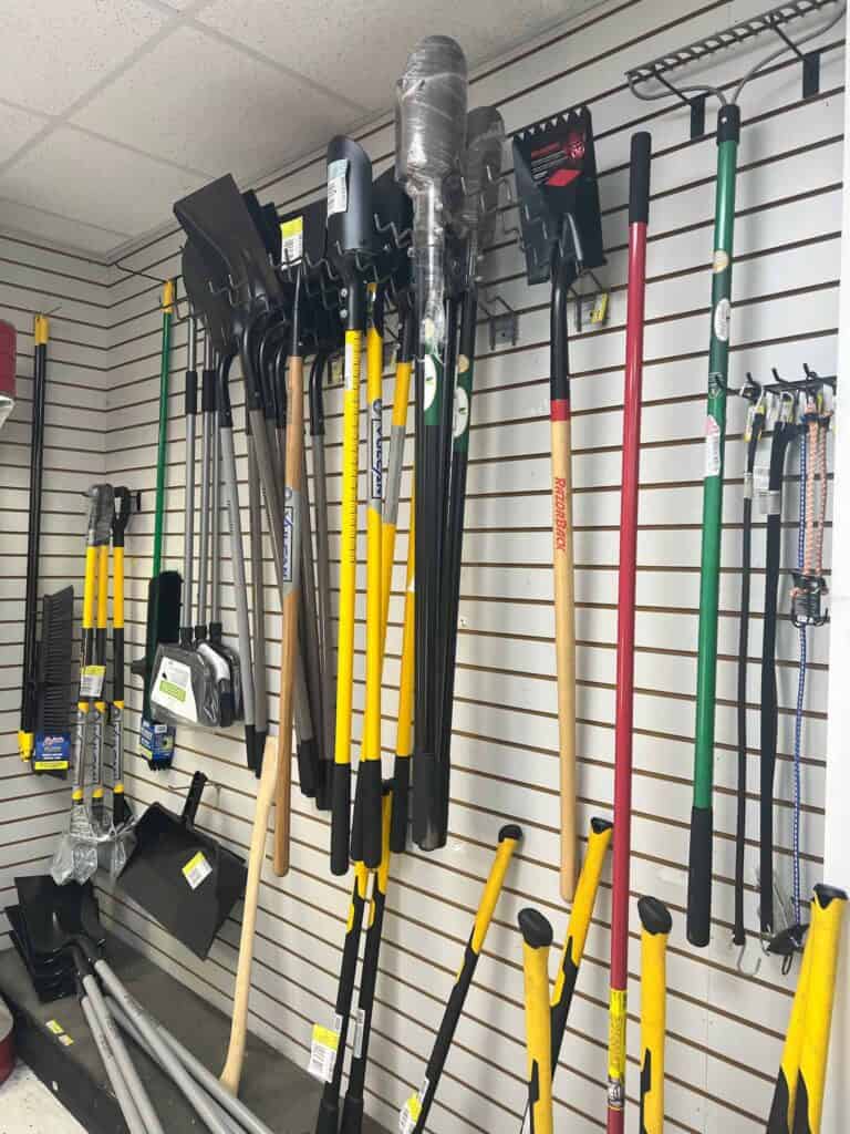 At Scott's Hill Hardware, a store display showcases various tools including shovels, axes, picks, and handles hanging neatly on a slatted wall. Prices and labels are visible on some items.