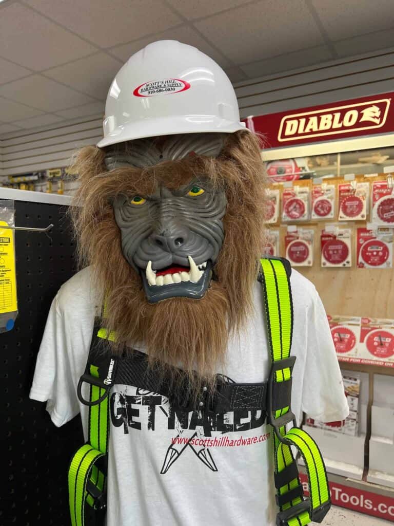 A mannequin at Scott's Hill Hardware sports a hard hat, a wolf mask, a safety vest, and a t-shirt that reads "www.scottshillhardware.com." Positioned in the store, it stands amidst an array of tool products in the background.