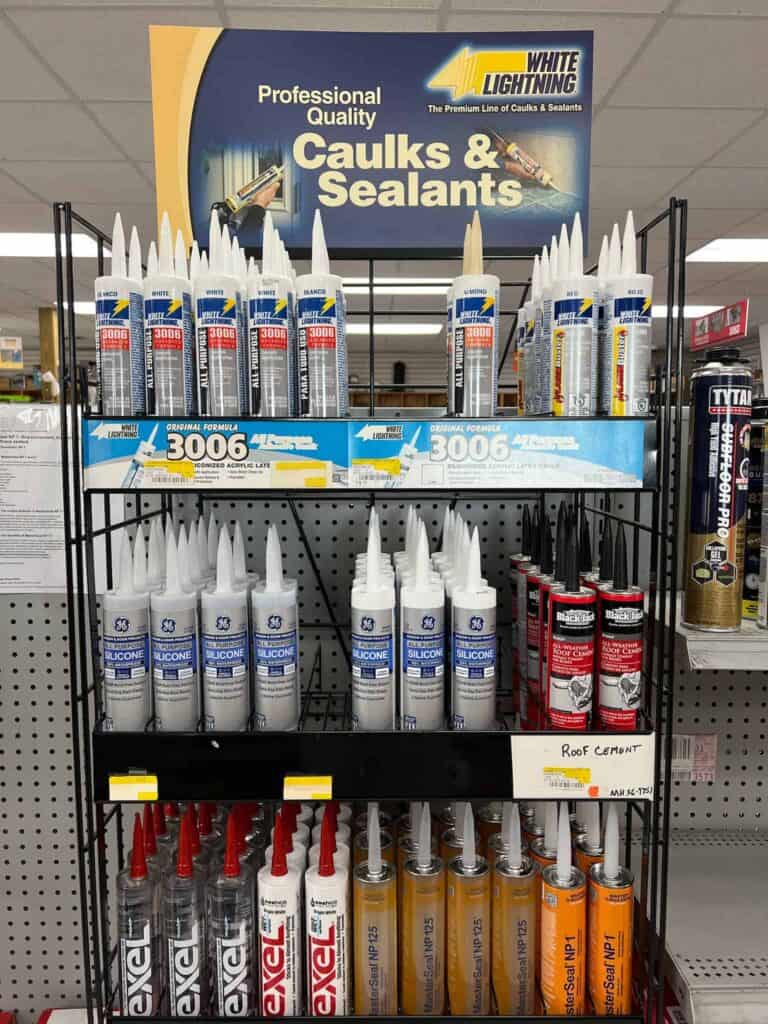 A store display rack at Scott's Hill Hardware showcasing various caulks and sealants, including brands like White Lightning, GE Silicone, and Excel. Products are arranged on three shelves with labels for easy identification.