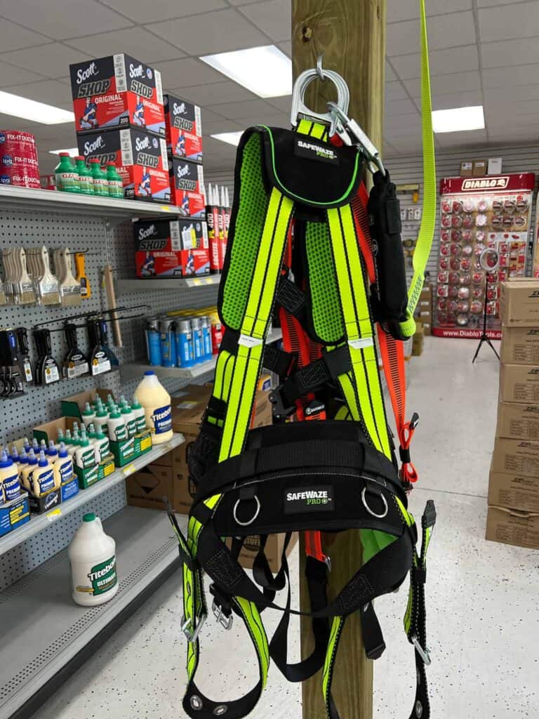 A neon green safety harness with black straps hangs on a wooden post at Scott's Hill Hardware, surrounded by various tools, paints, and hardware items on shelves.