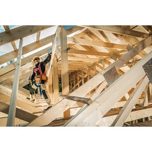 A person wearing construction gear stands on scaffolding within the wooden framework of a building under construction, surrounded by various beams and planks in daylight. Nearby, freshly delivered supplies from Scott's Hill Hardware are neatly stacked, ready for use.