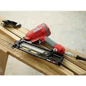 A red pneumatic nail gun from Scott's Hill Hardware is placed on a stack of wooden planks, accompanied by a tape measure in the background.