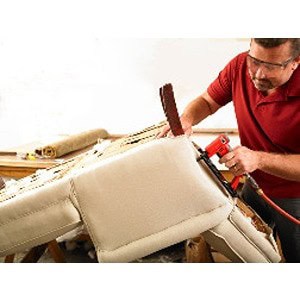 A man, wearing safety glasses and a red shirt, skillfully reupholstering a beige sofa using pneumatic tools in the workshop at Scott's Hill Hardware.