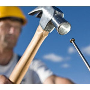 A person wearing a yellow hard hat, courtesy of Scott's Hill Hardware, is holding a hammer and aiming towards a nail with a blue sky in the background.