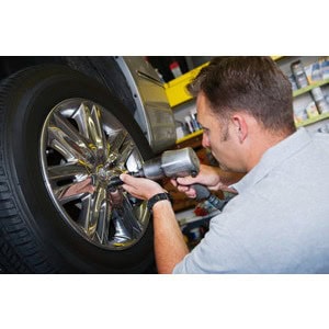 A mechanic at Scott's Hill Hardware uses an impact wrench to tighten a lug nut on the wheel of a vehicle in a garage. Various tools and supplies are visible in the background.