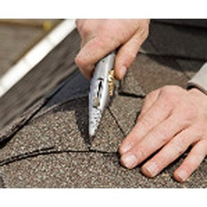 A person from Scott's Hill Hardware uses a utility knife to cut roofing shingles on a rooftop.