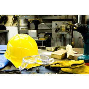 A yellow hard hat, safety goggles, gloves, and various tools from Scott's Hill Hardware are placed on a workbench in a workshop environment.