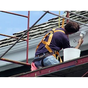 A person wearing a safety harness from Scott's Hill Hardware is painting the trim of a roof while standing on scaffolding.